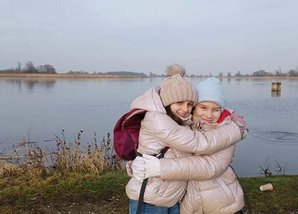 Na pierwszym planie dwie dziewczynki przytulaja się, w tle rzeka