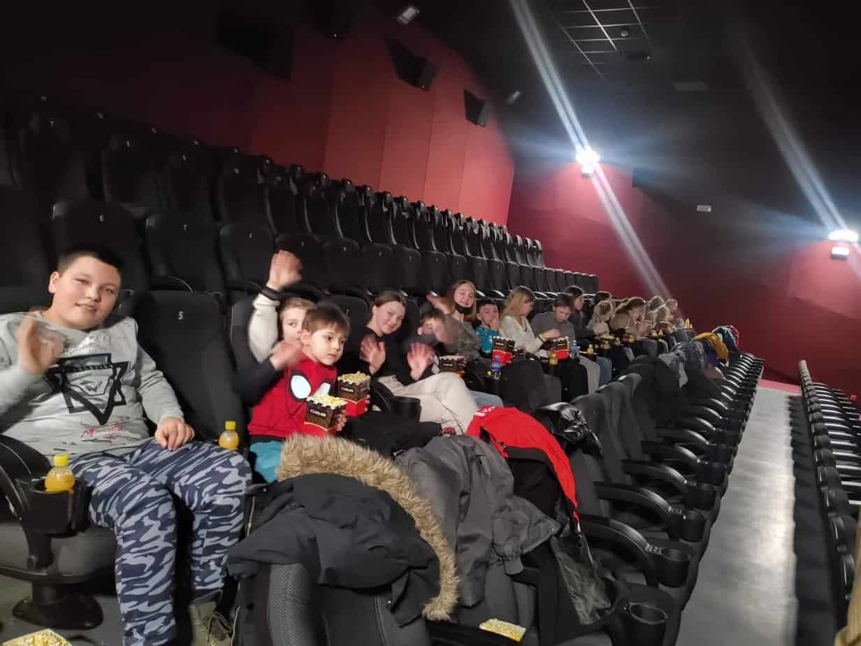 grupa dzieci siedzi w jednym rzędzie sali kinowej, inne rzędy puste, ściany ciemne
