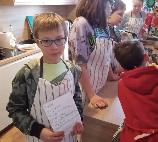 grupa dzieci ubranych w fartuszki, stoi w kuchni przy blacie kuchennym, chłopiec na pierwszym planie trzyma pokazuje przepis na ciasto