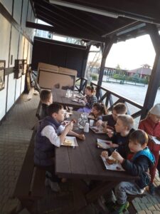 kilkoro dzieci siedzi pod wiatą, przy stołach, jedzą kiełbasy z grila