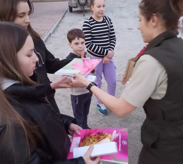 kobieta prowadząca zajęcia rozdaje dzieciom foldery papierowe, w tle przyczepka zielona