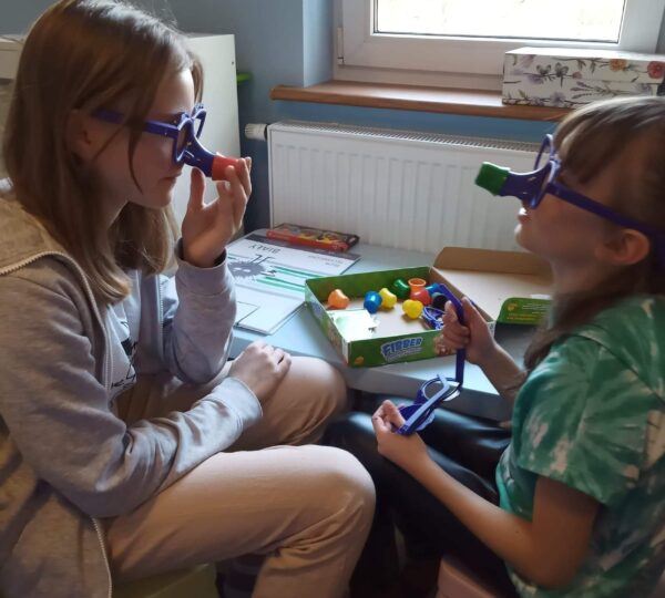 dwie dziewczynki bawią się przy stoliku. na nosach mają śmieszne okulary z wydłużonym nosem.