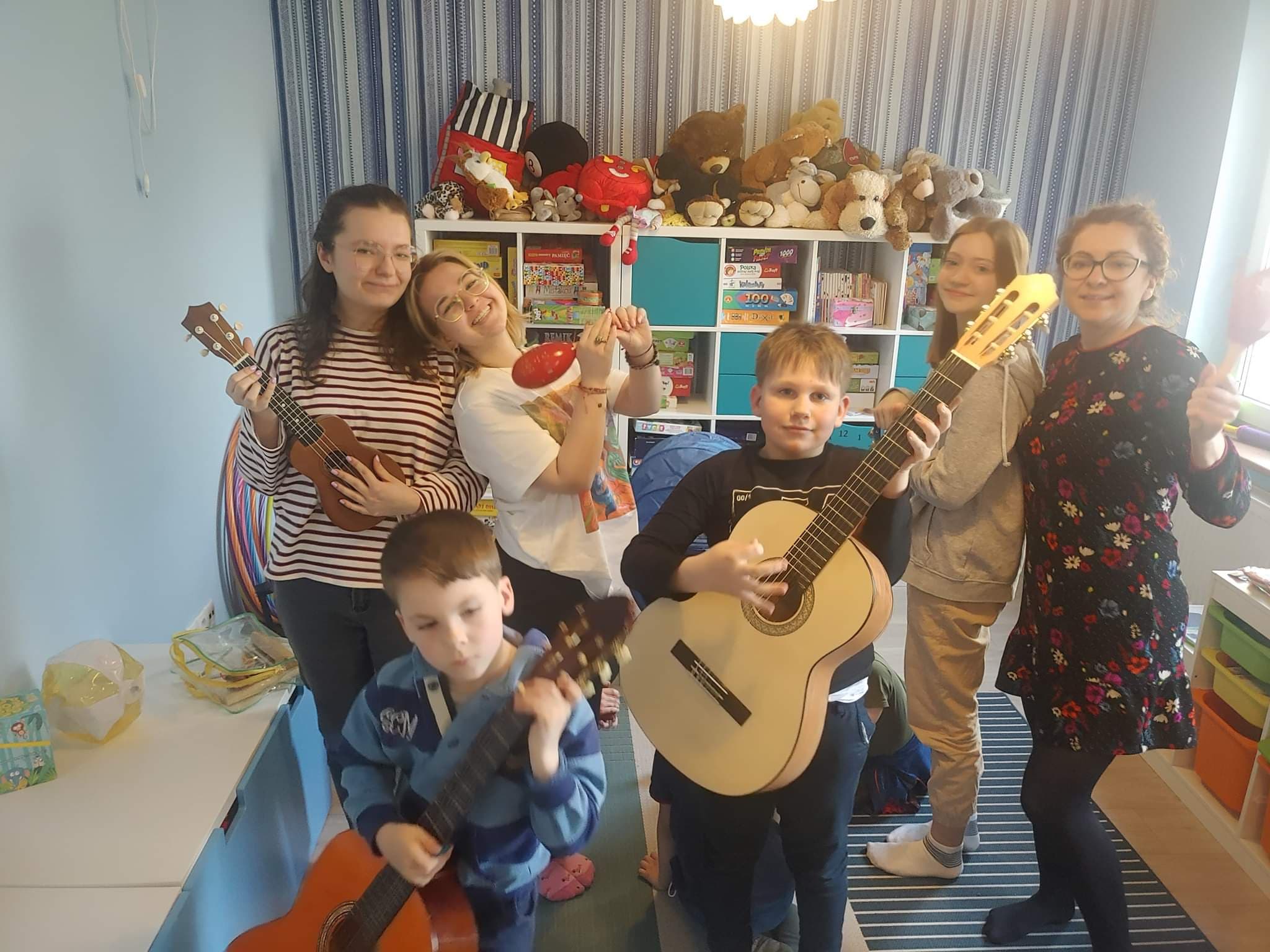 6 osób stoi w pomieszczeniu, dzieci trzymają instrumenty ( gitara, ukelele, marakas). W tle regał z zabawkami i kolorowa ściana.