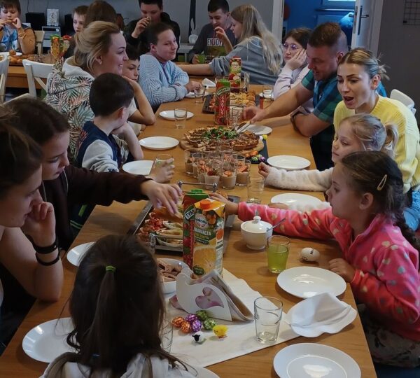 liczna grupa dzieci i dorosłych siedzi przy stole zastawionym poczęstunkiem