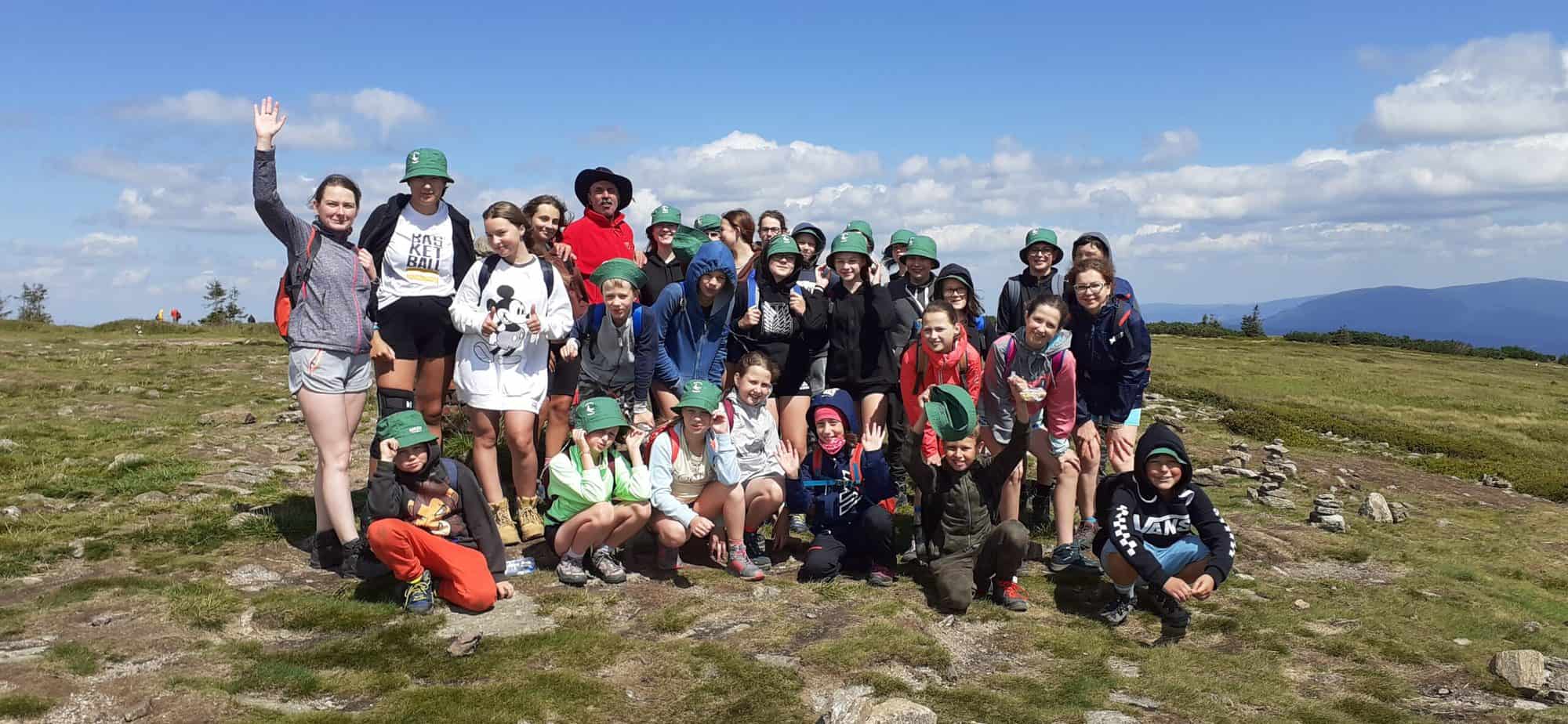 Grupa dzieci, młodzieży i dorosłych pozuje do zdjęcia na szczycie góry. w tle pasmo gór i błękitne niebo z chmurami.