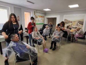 w pomieszczeniu seniorzy siedzą na krzesłach, ubrania zabezpieczone folią fryzjerską, uczennice robią seniorom fryzury