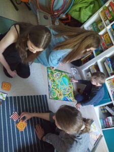 czworo dzieci siedzi na dywanie i gra w gry planszowe, w tle regał z książkami i grami
