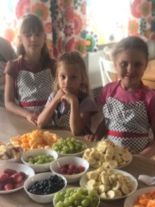 Trzy dziewczynki w fartuszkach w kropki stoją przy blacie kuchennym, przed nimi miski pełne pokrojonych owoców