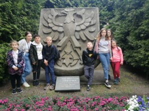 7 dzieci stoi przy dużej kamiennej rzeźbie z godłem Gorzowa, dokoła zieleń i kwiaty