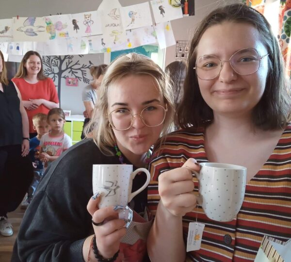 dwie dziewczyny okularach pozują do zdjęcia trzymając kubki z herbatą, w tle inne osoby oraz kolorowe pomieszczenie