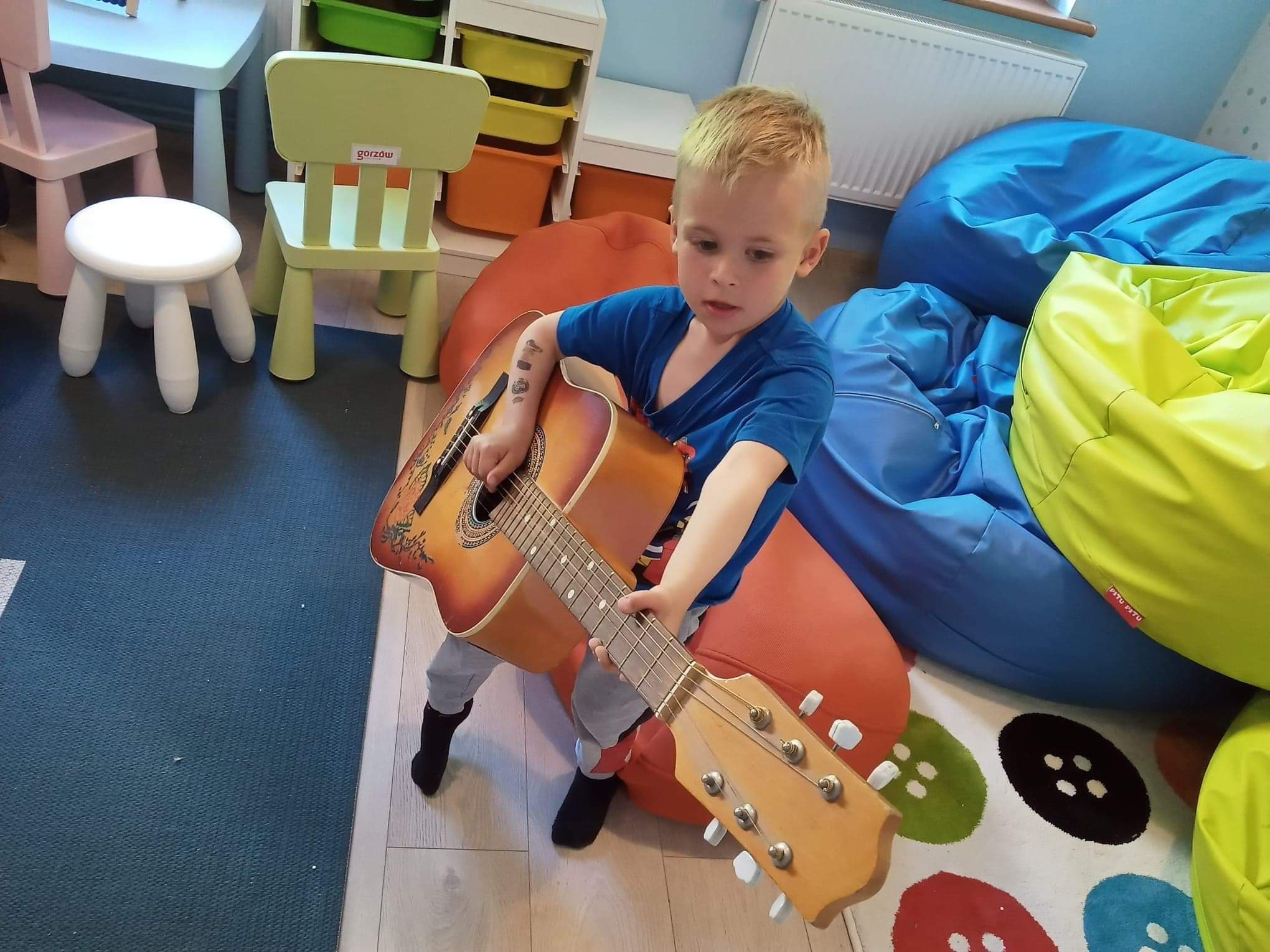 Chłopiec gra na gitarze, w tle kolorowe krzesełka i pufy, dywan