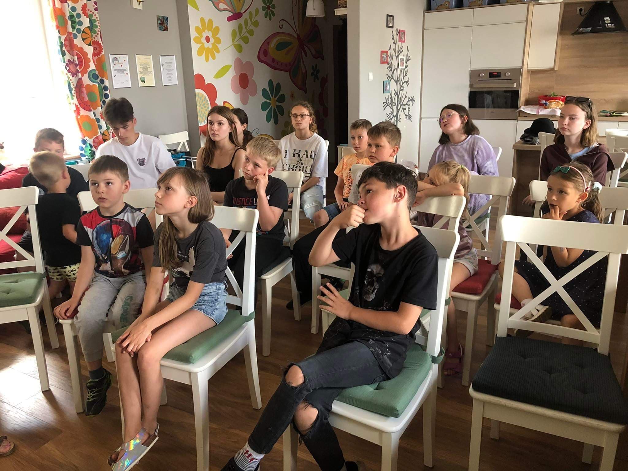 grupa dzieci siedzi na krzesłach, w pomieszczeniu, z uwagą słuchają kogoś kto przed nimi stoi