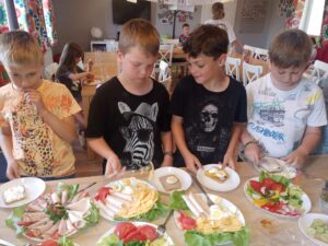 czterej chłopcy stoją przy blacie kuchennym, przed nimi talerze pełne warzyw, sera, jaj i wędliny