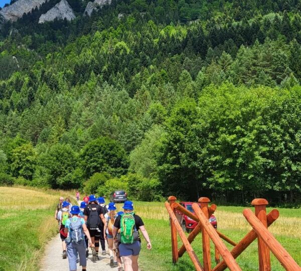grupa osób idzie szlakiem górskim, po prawej drewniane ogrodzenie, w tle szczyt