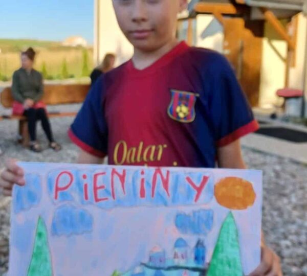 chłopiec stoi przed budynkiem, prezentuje obraz namalowany farbami, na nim napis "PIENINY"