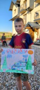 chłopiec stoi przed budynkiem, prezentuje obraz namalowany farbami, na nim napis "PIENINY"
