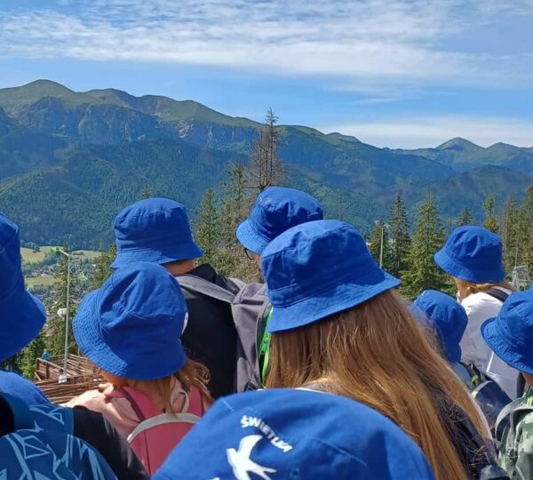 grupa osób w niebieskich kapelusikach, odwórcona tyłem do fotografa patrzy na pasmo górskie