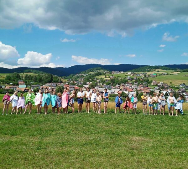 liczna grupa dzieci stoi w rzędzie, wszyscy otuleni recznikami kąpielowymi, w tle miasteczko i góry