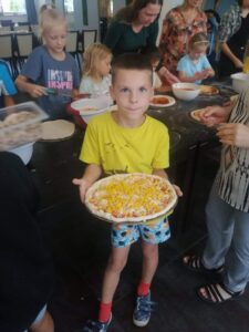 chłopiec prezentuje surową pizze, udekorowaną sosem, serem i kukurydzą, w tle inne dzieci dekoruja swoje pizze
