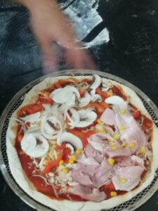 pizza przed upieczeniem, na cieście leżą kawałki pieczarek, szynki, cebuli, papryki