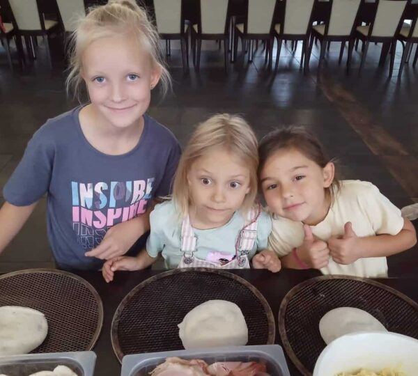 trzy dziewczynki stoja przy stole, przed nimi stoja koszyki z wyrobionym ciastem na pizze