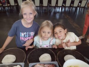 trzy dziewczynki stoja przy stole, przed nimi stoja koszyki z wyrobionym ciastem na pizze