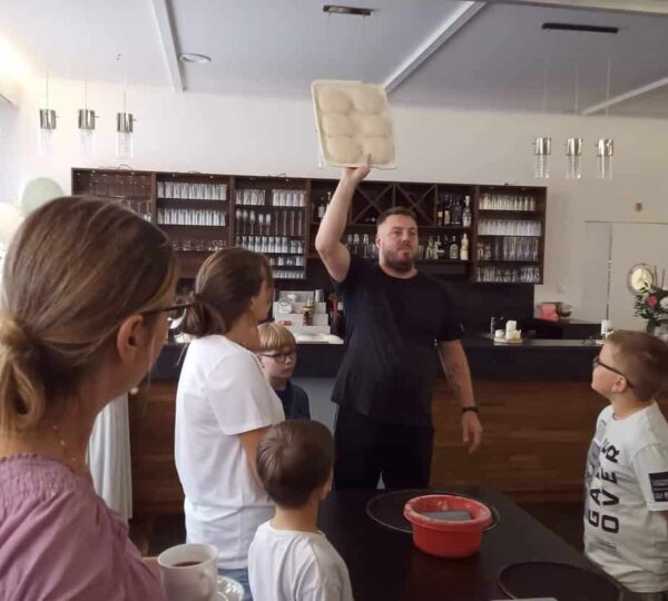 uczestnicy zajęć patrzą na kucharza, który trzyma wysoko uniesioną blachę z wyrobionym ciastem do pizzy