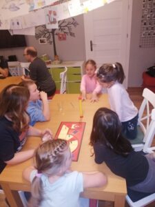 Grupa dzieci zgromadzona przy stole, gra w grę planszową, w tle inne osoby grające w inne gry