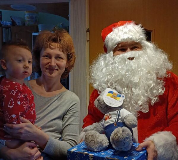 Kobieta z chłopczykiem na rękach pozuje obok Mikołaja, Mikołaj trzyma w dłoniach zapakowany prezent oraz misia.