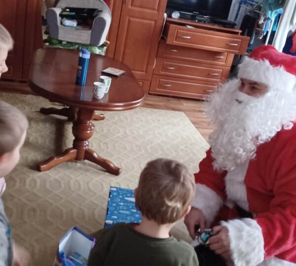 Sytuacja w pokoju umeblowanym w kolorach brązu i beżu. Mikołaj kuca przed trojgiem małych dzieci i wrecza im upominki.