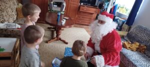 Sytuacja w pokoju umeblowanym w kolorach brązu i beżu. Mikołaj kuca przed trojgiem małych dzieci i wrecza im upominki.