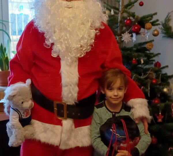 "Mikołaj" pozuje do zdjęcia z chłopcem, który siedzi na specjalistycznym fotelu. Chłopiec uśmiecha się, trzyma na kolanach prezent, a na głowie ma czapkę mikołajową