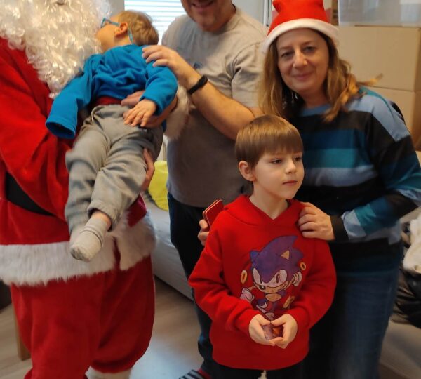 Mikołaj trzyma na rękach dziecko, obok stoi tata, pani psycholog z hospicjum oraz chłopczyk w czerwonej bluzie.
