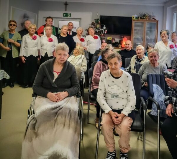 liczna grupa osób pozuje do zdjęcia, z przodu pacjenci i seniorzy, z tyłu stoja członkowie zespołu muzycznego w białych koszulach z czerwonym kwiatem